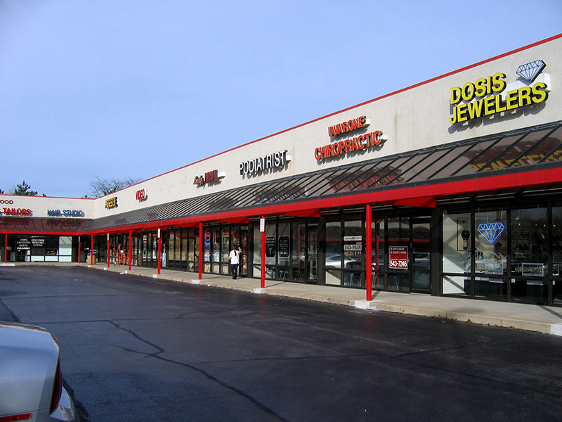A small strip mall with a row of storefronts under a flat roof and a red awning supported by red poles. Business signs include a jeweler, podiatrist, chiropractic office, hair studio, and other local shops. The storefronts have large glass windows, and a few people are seen walking near the entrances. The parking lot in front is mostly empty, with only one visible car. The sky is clear and blue.