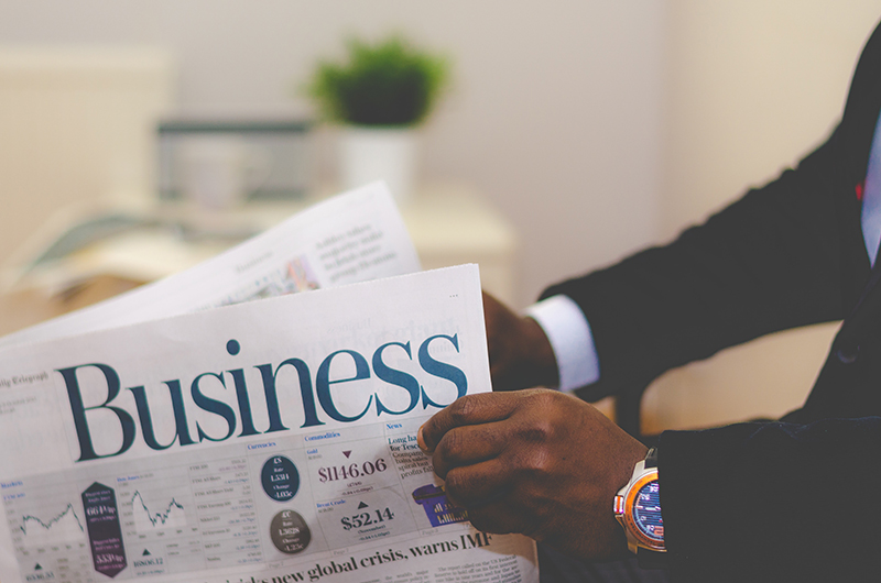 A person wearing a dark suit and a colorful wristwatch is holding a newspaper open to the Business section. The newspaper displays stock market information, graphs, and headlines. A blurred background shows a white desk with a potted plant and an open laptop.