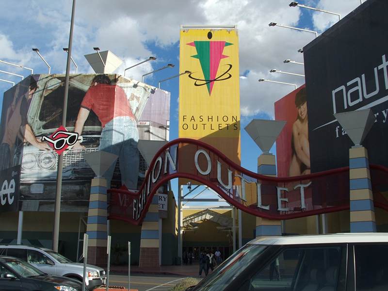 The entrance to a Fashion Outlets shopping center on a partly cloudy day. Large colorful banners and advertisements for various brands, including Lee and Nautica, decorate the front of the building. A tall yellow banner in the center reads “Fashion Outlets” with a stylized logo above it. A red, wavy metal arch spelling “FASHION OUTLET” spans across the entrance. Several cars are parked in the foreground, and people can be seen walking into the mall.