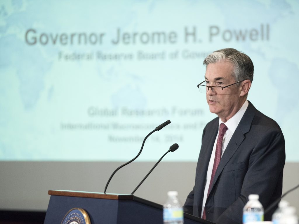 Photo of Jerome Powell standing at a podium with a powerpoint shown on a screen behind him.