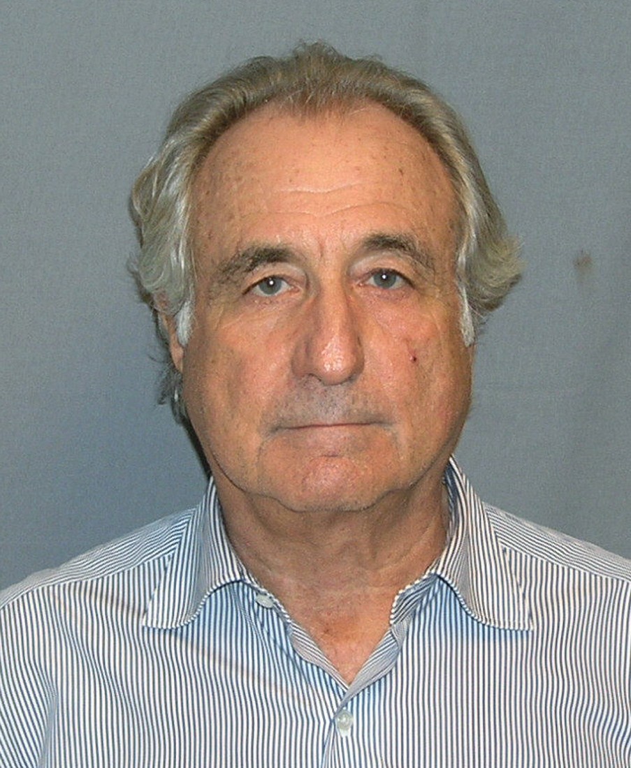  A photograph of Bernie Madoff standing in front of a blue background. He's an older white man with white hair and a flat smile. He's wearing a striped collared shirt.