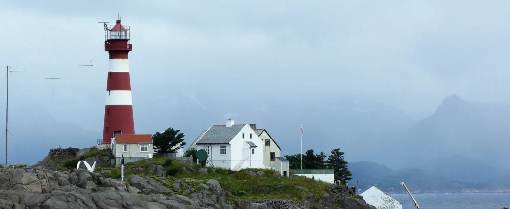 Photo of a lighthouse and other building with a backdrop of water and mountains.