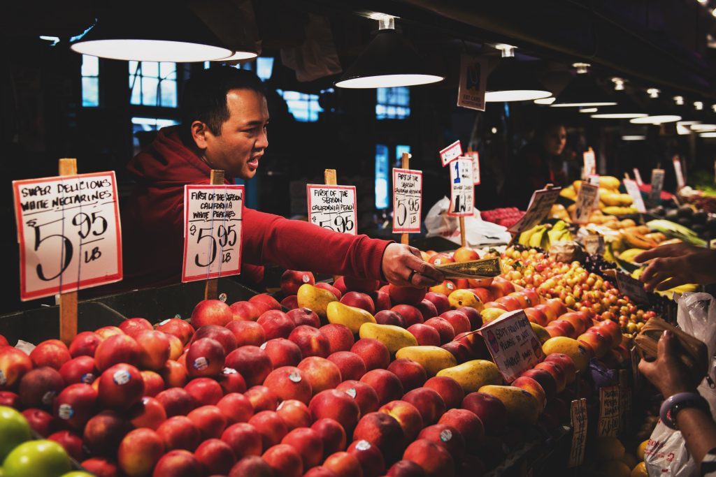 Photo of a man selling fruit to an unseen person. Several stands with fruit are in front of the man. 